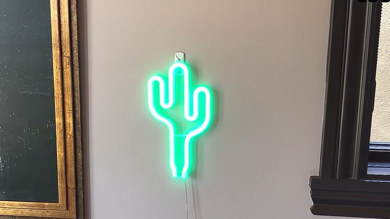 Cactus-shaped neon sign hanging on a wall next to a framed print and a window