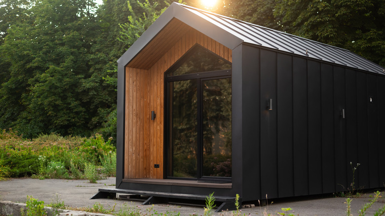 A modern tiny house made of black wood with large glass doors in front of a forest