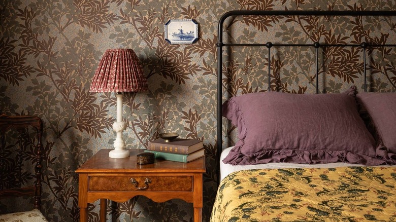 Old English cottage style bedroom with dark wallpapered walls, black wire bed frame, yellow bedspread, and wooden night table.