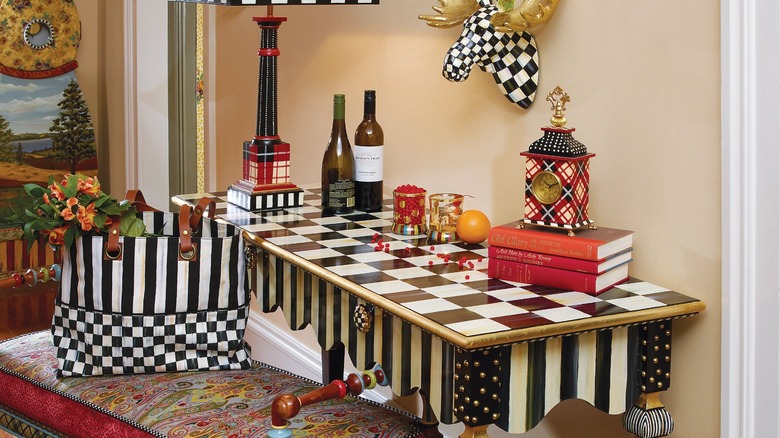 MacKenzie-Childs console in courtly check pattern topped with books, bottles, and a clock, next to a colorful and intricate bench