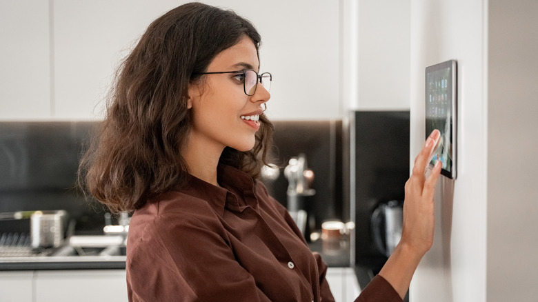 A smiling woman uses smart home interface mounted on a kitchen wall.