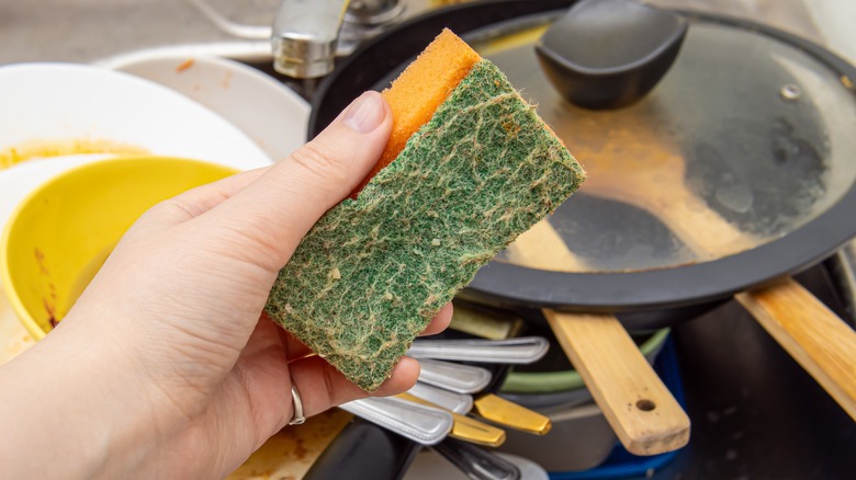 Hand holding dirty orange and green sponge with dirty dishes in the background