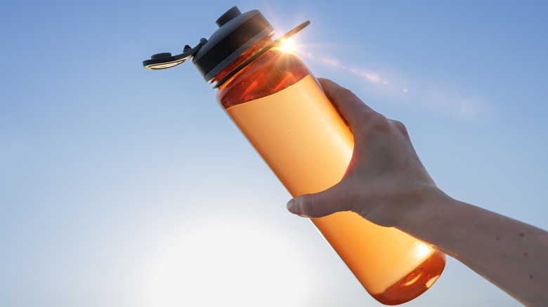 Close-up of a woman's hand holding an orange plastic water bottle against a blue sky background