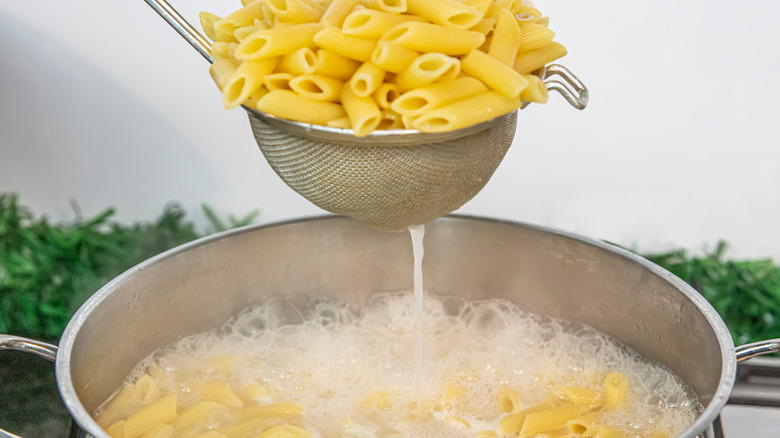 A small sieve holding cooked penne pasta above a boiling pot of water and pasta.
