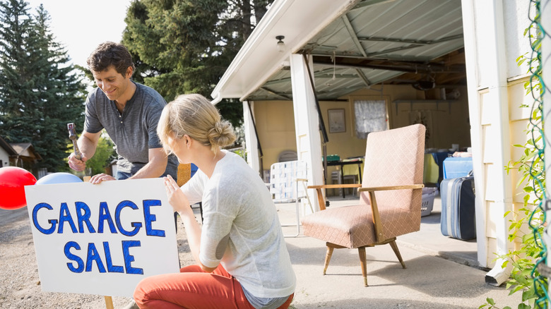 Man and woman installing garage sale sign outside their garage, with chairs in background
