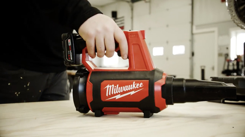 A person holding Milwaukee's M12 Lithium-Ion Cordless Compact Spot Blower on a table in a workshop