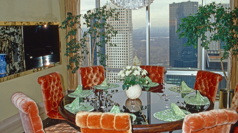 A shiny round dining table with a glass top surrounded by orange crushed velvet chairs