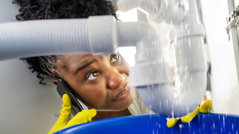 A woman is on the phone while dealing with a leaking pipe under a sink
