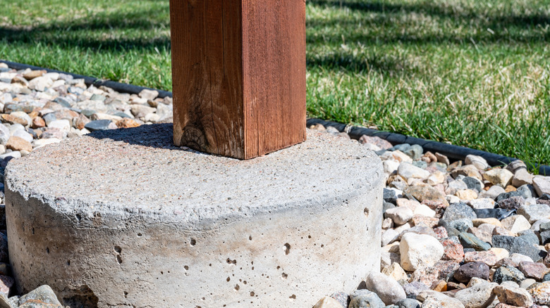 A wooden post is embedded in a concrete footer that sits atop gravel