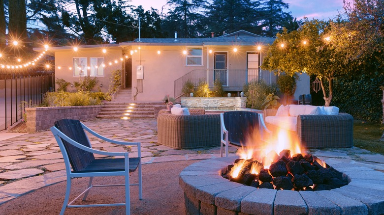 A backyard fire pit surrounded by chairs and other decor with twinkling lights at dusk