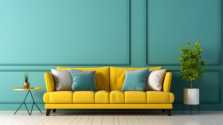 A teal wall with a bright yellow sofa with teal, grey and white cushions. A house plant sits beside it in a pot.