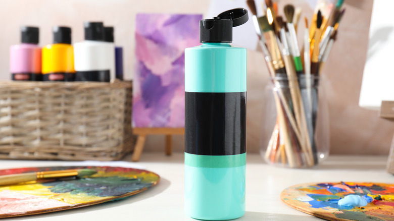 An open bottle of acrylic paint sits on a desk with other painting supplies in the background.