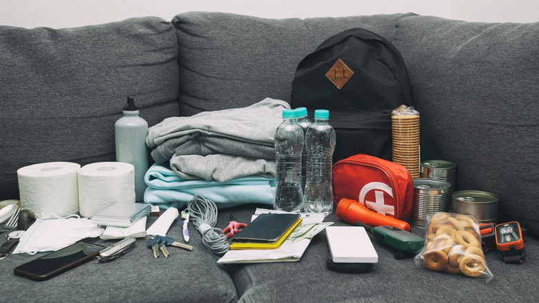 emergency kit with blankets, food, water, toilet paper, and other supplies laid out on a gray couch
