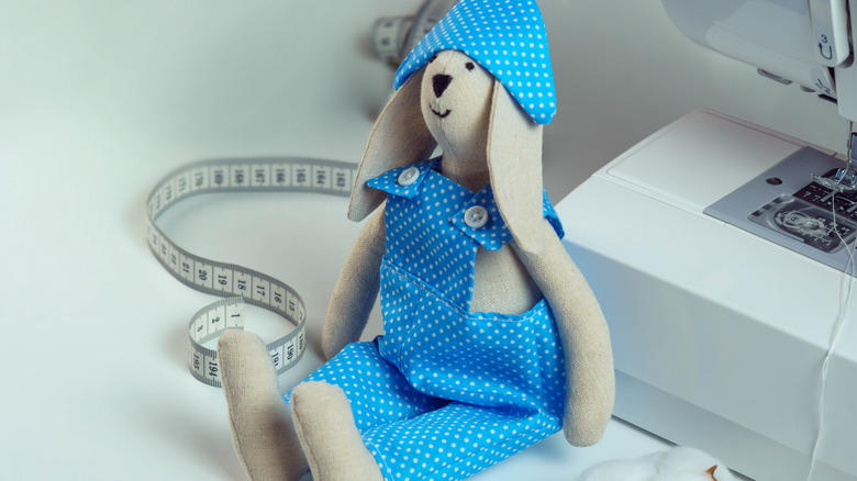 A smiling homemade stuffed animal bunny in cloth overalls next to a sewing machine with measuring tape