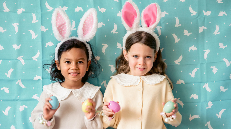 two little girls in bunny ears holding easter eggs in front of bunny sheet backdrop