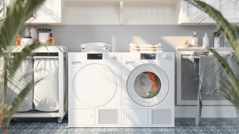 A white front-load washer next to a matching dryer in a neutral-toned laundry room with plants