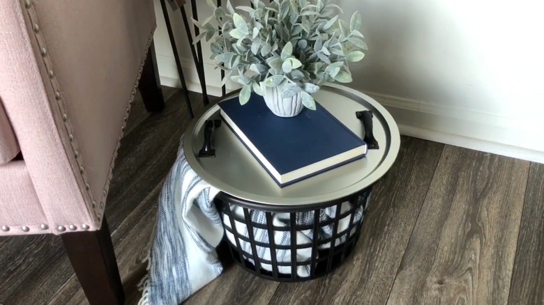 A black Dollar Tree laundry basket with a blanket inside and a round silver tray on top as a table surface.