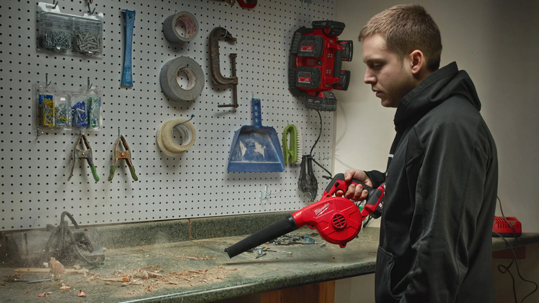 A man using Milwaukee's M18 18V Lithium-Ion Cordless Compact Blower to clear a dirty workbench