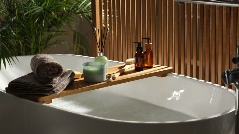 Wooden bath tray with candle, air freshener and bathroom amenities on tub indoors