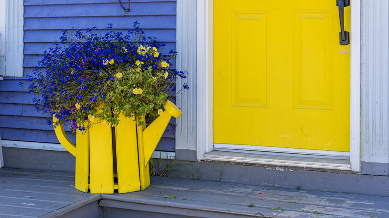 Bright yellow watering can flower pot sitting on a front porch with a yellow door