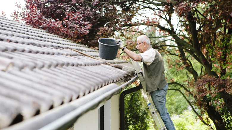 An older man standing on a ladder and cleaning out the gutters.