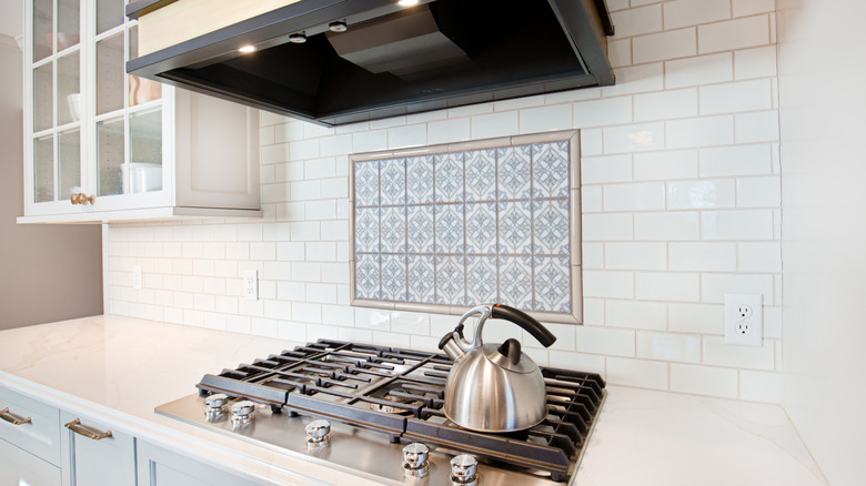 A white kitchen with a stove and a subway tile backsplash with a painted tile accent in the center.