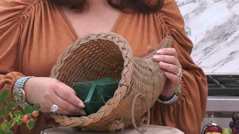 A woman wearing lots of jewelry pushing a piece of floral foam into a wicker cornucopia