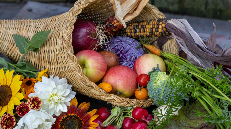 Wicker cornucopia filled with apples, colorful corn, and other produce with fall flowers next to it