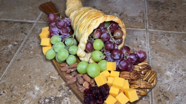 A bread cornucopia on a cutting board stuffed with red and green grapes and surrounded by cheese, nuts, and dried fruits