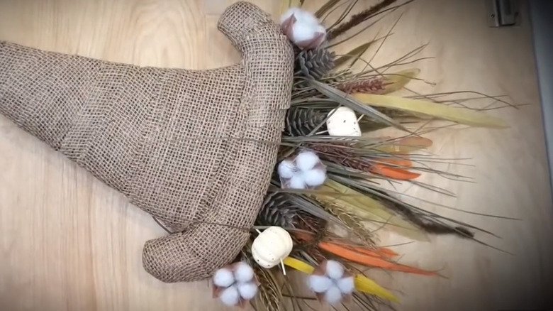 A fabric cornucopia filled with wheat, plants, cotton, pinecones, and small white pumpkins