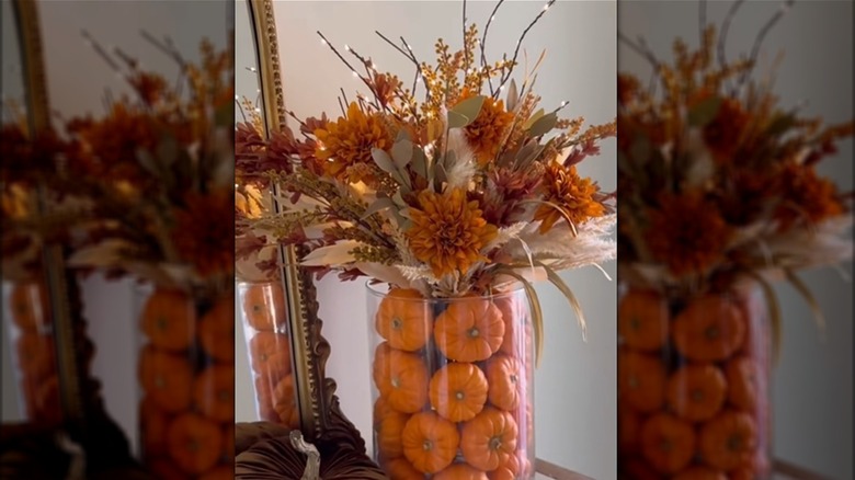 A glass vase is filled with small pumpkins and another vase full of flowers.