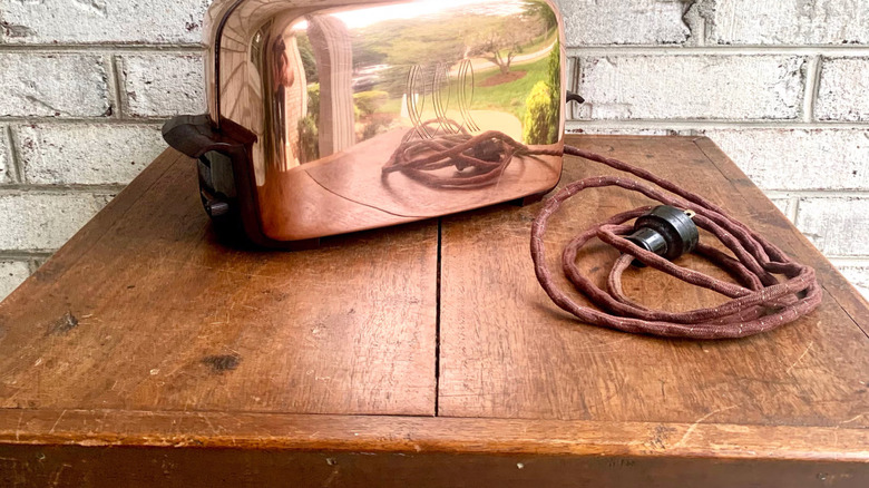 A vintage copper toaster with intact cord sits on a wooden table in front of a brick wall