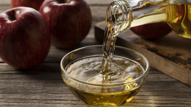 Apple cider vinegar being poured into a small clear glass bowl with apples on a wooden surface in the background
