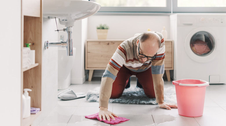 A man drying a bathroom floor beneath a leaking sink with a cloth