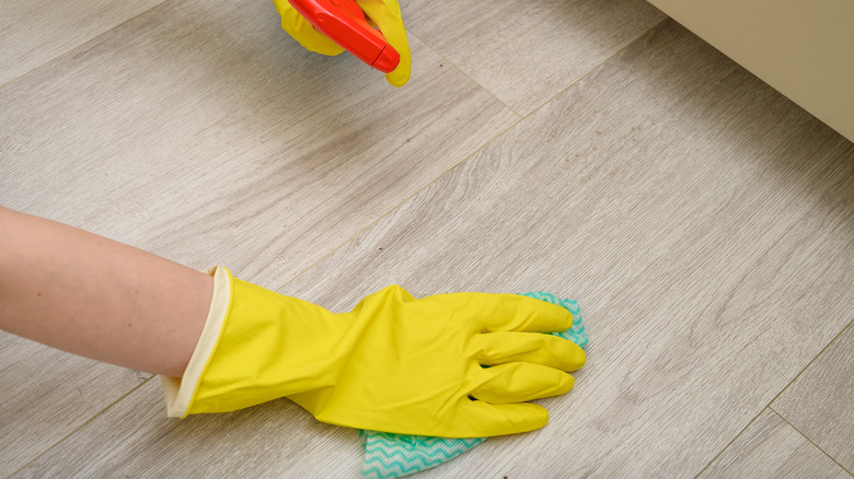 Hands with yellow plastic gloves hold a cloth and a spray bottle while washing the floor