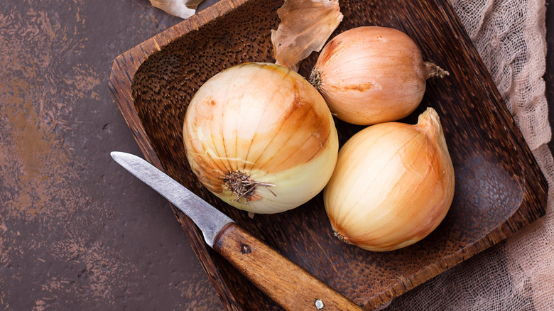 A wooden tray of onions and a knife are set on a rusted surface