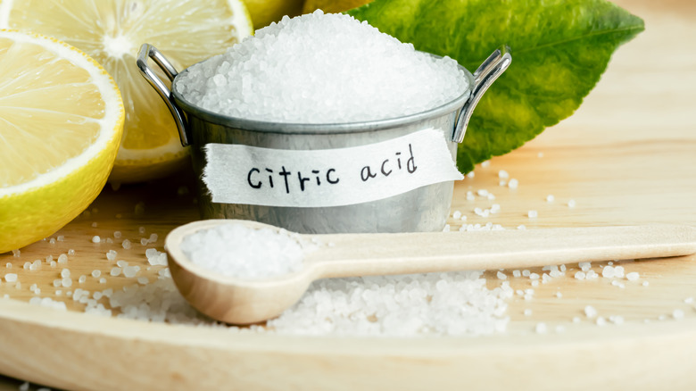 A heaping silver measuring cup labeled "citric acid" sits in front of lemons and behind a wooden scoop with more citric acid