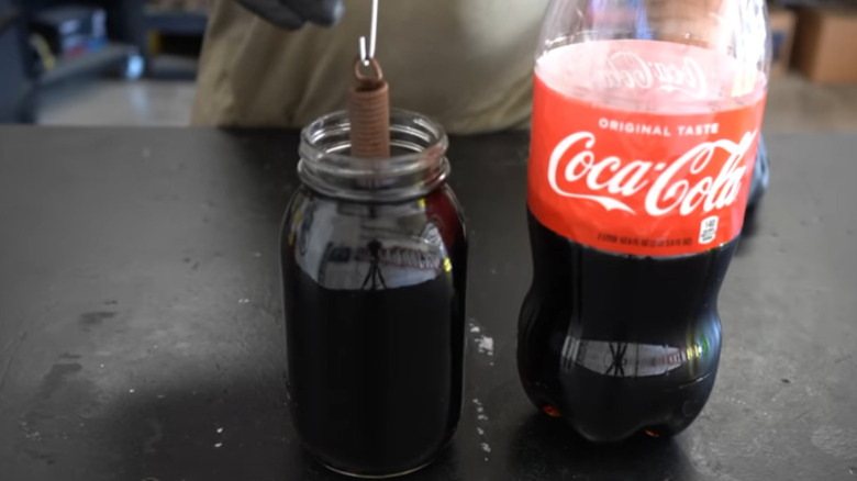 A rusted coil is being lowered into a jar of cola, next to a bottle of Coca-Cola