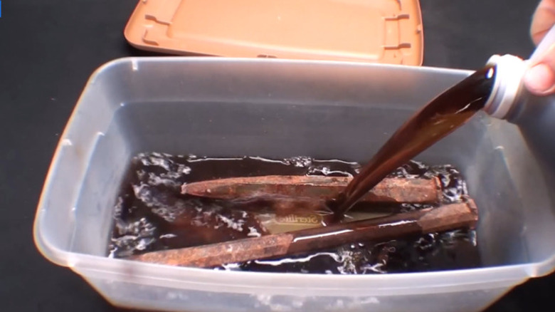 A hand is pouring a bottle filled with diluted molasses into a clear container holding rusty tools