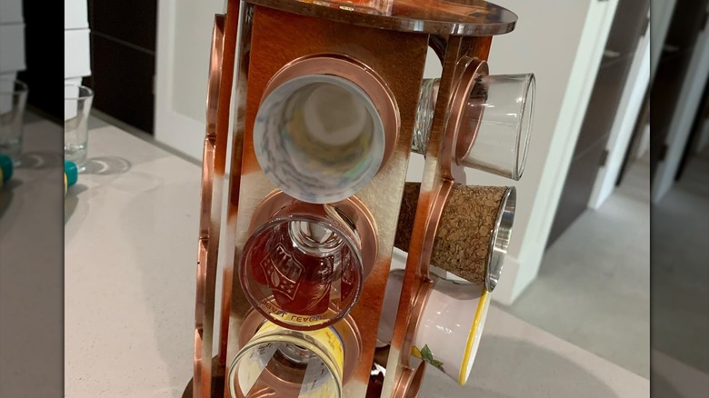 A brown wooden Keurig K-cup holder holding various decorative shot glasses on a counter