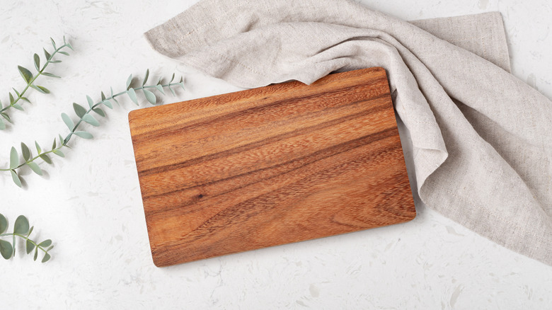 Wooden cutting board sitting on counter with dishtowel and Eucalyptus stems next to it.