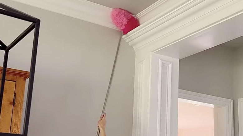 A homeowner cleans crown molding with a pink long-handled mop.