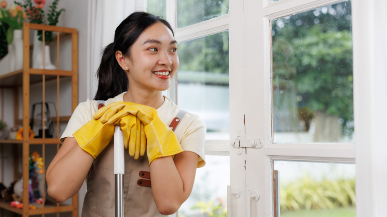 A woman wearing gloves looks out a clean window while leaning on a mop handle.