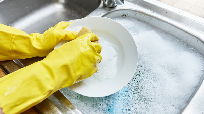 Close-up of gloved hands washing dishes in a sink full of soapy water
