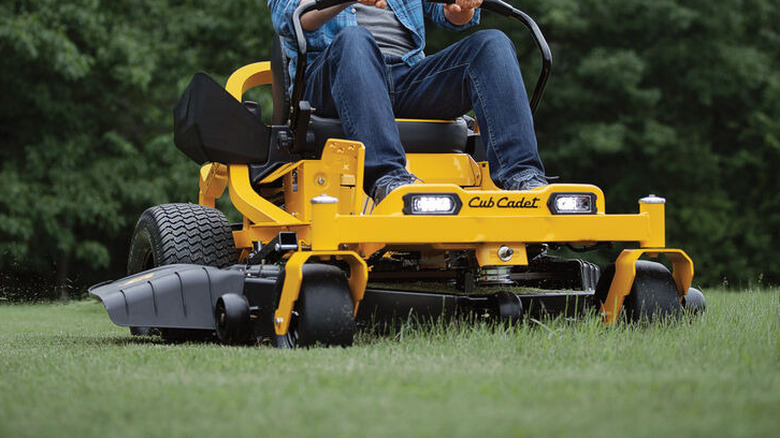 Man steering yellow Cub Cadet mower across a large lawn with tree backdrop