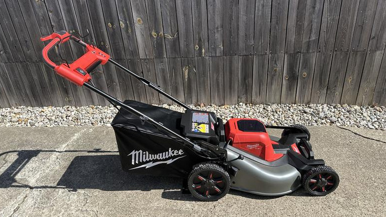 Silver and red Milwaukee push mower on a pavement by gravel and a wood fence