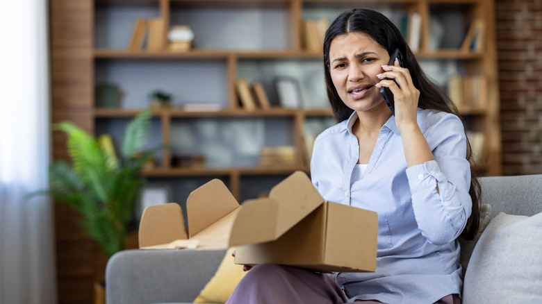 A frustrated woman talking on the phone in her living room, with a cardboard box in her lap.