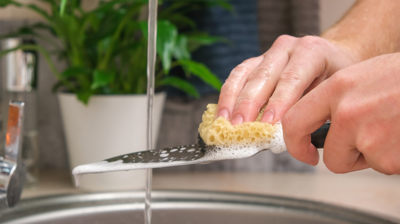Hands using sponge to wash large kitchen knife in sink