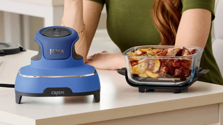 The blue Ninja Crispi air fryer next to a glass container of food.