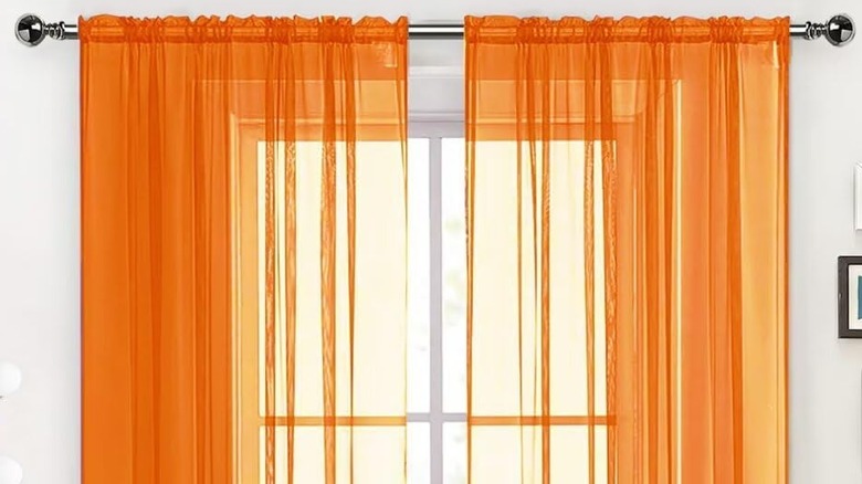 A white wall around a window dressed with a sheer orange curtain.